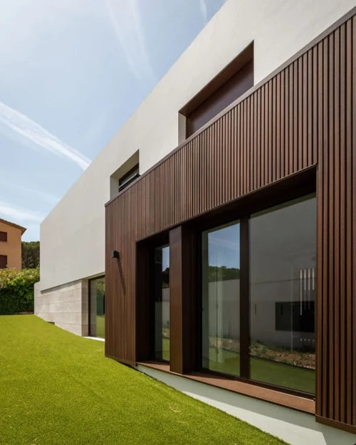 Fachada de casa moderna con revestimiento de listones de madera oscura, ventanales y pasto